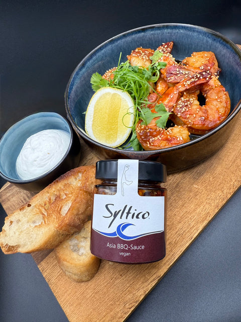 Garnelen in roter Asia BBQ Marinade mit Sesam und einem Zitronentück. Die Garnelen sind ein einer dunkelblauen Schale welche auf einem Holzbett steht. mit auf dem Brett eine kleine Schale mit Aioli und 2 Scheiben geröstetes Baguette. An das Brett angelegt ist ein Glas der Marke Syltico mit einer Asia BBQ Sauce und der Insel Sylt mit auf dem Etikett