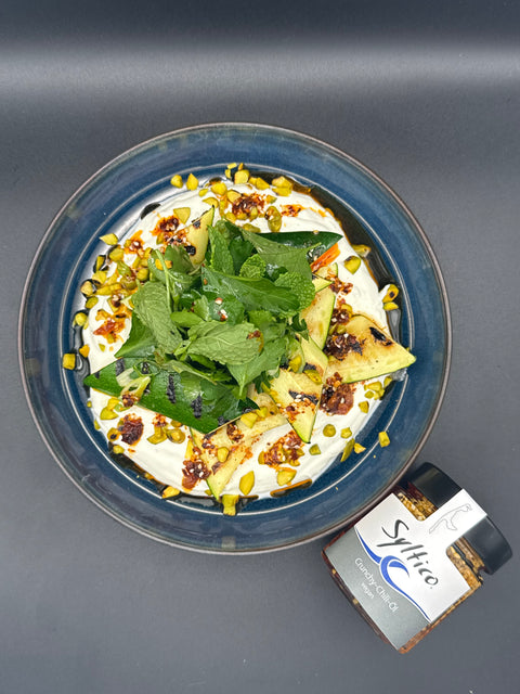 Ein schmackhaft aussehender Salat auf einem dunkel blauen Teller. Vor dem Teller lliegt ein Glas Crunchy Chili Öl der Marke Syltico mit der insel Sylt auf dem Etikett. Der Salat besteht aus einer Joghurt Creme die den Teller gestrichen wurde getopt von gegrillten Zucchini, gerösteten Pistazien unnd einem Salat auf Minze und Petersilie. das Crunchy Chili öl wurde ansprechend auf den Salat geträufelt 