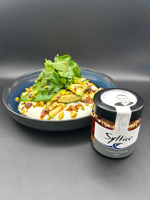 Ein schmackhaft aussehender Salat auf einem dunkel blauen Teller. Vor dem Teller steht ein Glas Crunchy Chili Öl der Marke Syltico mit der insel Sylt auf dem Etikett sowie dem Portrait des Küchenmeisters Nicolas Kluth auf dem Deckel. Der Salat besteht aus einer Joghurt Creme die den Teller gestrichen wurde getopt von gegrillten Zucchini, gerösteten Pistazien unnd einem Salat auf Minze und Petersilie. das Crunchy Chili öl wurde ansprechend auf den Salat geträufelt 
