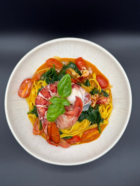 Auf einem weißen Teller angerichtet gelbe Linguinie Pasta mit in einer Hummerpastasauce sowie einem aufgelösten Hummerschwanz und einer aufgelösten Hummerschere die Pasta wurde verfeiner mit Kirschtomaten, frischem Blattspinat und Basilikum 