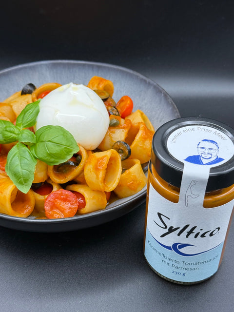 Runde Ringförmige Pasta mit Karamellisierter Tomtenauce auf einem Schwarzen Teller mit Kirchtomaten, Oliven, Basilikum und einem Burrata. ein Glas von der Syltico Kramellisierten Tomtensauce mit Parmesan und auf dem Deckel des Glases ist ein Portrait von dem Küchenmeister Nicolas Kluth