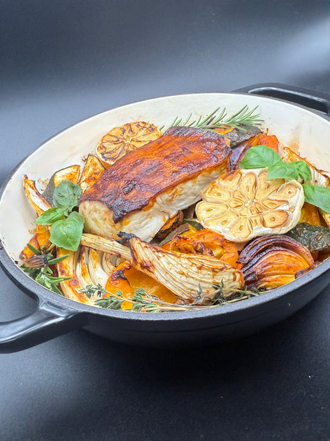 In einer Pfanne ist ein Stück Fisch, in dem Fall Heilbutt umgeben von geröstetem Gemüse Zucchini, Paprika, Karotten, Süßkartoffel und Fenchel mit einer gerösteten Zitrone und Knoblauch. mit in der Pfanne sind Rosmarin, Thymian und Basilikum. 