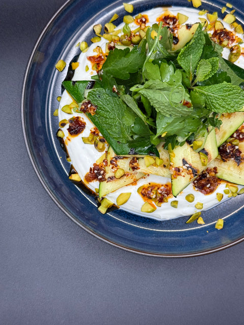 Ein schmackhaft aussehender Salat auf einem dunkel blauen Teller.  Der Salat besteht aus einer Joghurt Creme die den Teller gestrichen wurde getopt von gegrillten Zucchini, gerösteten Pistazien unnd einem Salat auf Minze und Petersilie. das Crunchy Chili öl wurde ansprechend auf den Salat geträufelt 