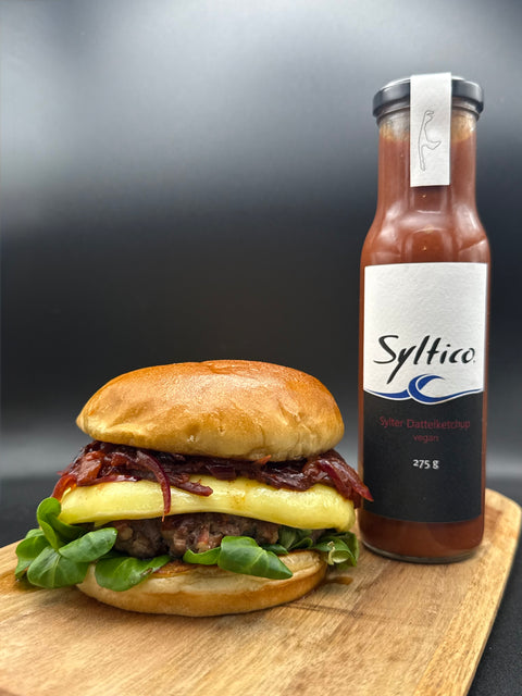 Auf eine Holzbrett steht ein Burger. Ein der Lammburger mit Feldsalat, Dattelketchup, geschmorten Zwiebeln, geschmolzenem Brie und wird gehalten von einem fluffigen Burgerbun. Nebem dem Burger Steht eine Glasflasche der Marke Syltico mit der Aufschrift Sylter Dattelketchup Vegan, ebenfalls auf dem Etikett sind die Umrisse der Insel Sylt. 