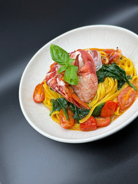 Auf einem weißen Teller angerichtet gelbe Linguinie Pasta mit in einer Hummerpastasauce sowie einem aufgelösten Hummerschwanz und einer aufgelösten Hummerschere die Pasta wurde verfeiner mit Kirschtomaten, frischem Blattspinat und Basilikum 