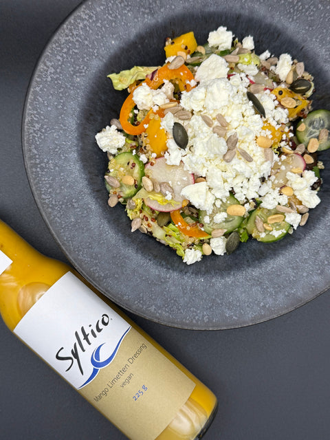 Appetitlich angerichteter  Salat auf schwarzem Teller und einer Glas Flasche mit gelbem Inhalt auf dem Etikett steht Syltico Mango Limetten Dressing der Salat besteht Römersalat, Quinoa, Gurken, Feta, Paprika. Körnern und Mango Limette Dressing  