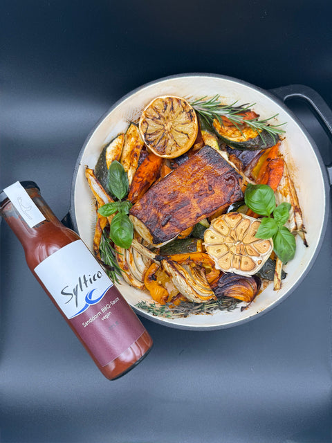 In einer Pfanne ist ein Stück Fisch, in dem Fall Heilbutt umgeben von geröstetem Gemüse Zucchini, Paprika, Karotten, Süßkartoffel und Fenchel mit einer gerösteten Zitrone und Knoblauch. mit in der Pfanne sind Rosmarin, Thymian und Basilikum. An die Pfanne angelehnt ist eine Flasche der marke Syltico mit einer Sanddorn BBQ Sauce.