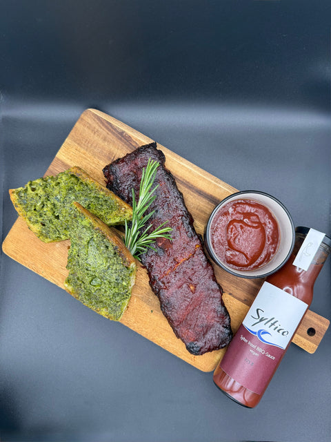 Spare Ribs auf einem Holzbrett angerichtet mit frisch geröstetem Knoblauchbaguett und einer günen Knoblauchcreme. Einem Zweig Rosmarin und so wie einer Dipschale in der Syltico BBQ abgefüllt ist. Am Rand von Holzbrett liegt eine Glas Flasche der Marke Syltico mit der Aufschrift Sylter ÖInsel BBQ Sauce.