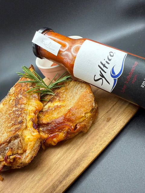 Auf einem Holzbrett sind 2 Grilled Cheese Sandwich übereinander angerichtet. Aus dem Sandwich läuft der geschmolzene Käse mit schmorten Zwiebeln. Angelehnt an eine kleine Schale mit Trüffelketchup ist eine Glasflasche der Marke Syltico mit der Aufschrift Sylter Trüffelketchup