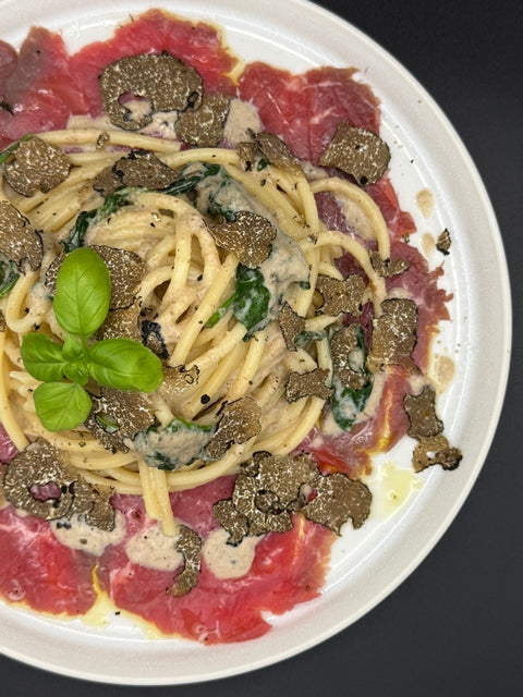 Spagetti mit Trüffelpastasauce liebevoll angerichtetet auf einem weißen Teller Die Pasta ist auf einem rotem  Rindercapaccio mit etwas Olivenöl angerichtet. Die Pasta wurde verfeinert mit frischem günen Blattspinat und frisch gehobelte Trüffelscheiben. 