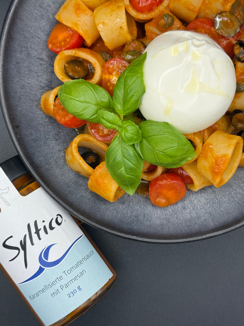 Runde ringförmige Pasta mit Karamellisierter Tomtenauce auf einem Schwarzen Teller mit Kirchtomaten, Oliven, Basilikum und einem Burrata. Ein liegendes Glas von der Syltico Kramellisierten Tomtensauce mit Parmesan 