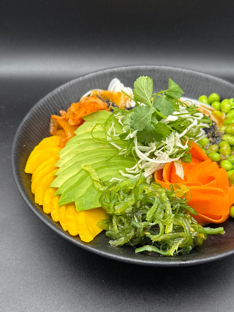 Sehr bunt angerichteter asiaticher Salat auf einem schwarzen Teller. Der Salat besteht aus eingerollten Glasnudel, die mit einem gerösteten Sesamdressing mariniert sind. auf dem Teller sind ebenfalls gefächerte Mango und Avocado sowie Karotten in Streifen schnitten, Edame, Kimchi, Wakame Algen und etwas Koriander und Frühlinglauch. 