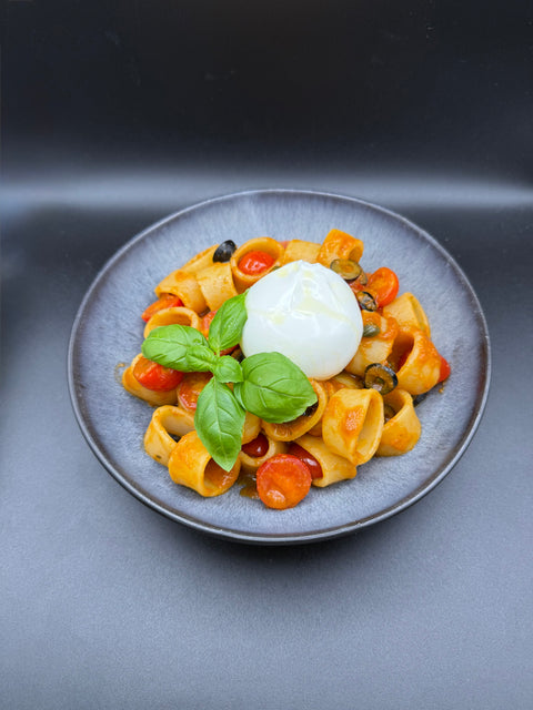 Appetitlich angerichtete Ringförmige  Pasta mit Karamellisierter Tomtenauce auf einem Schwarzen Teller mit Kirchtomaten, Oliven Basilikum und einem Burrata. 